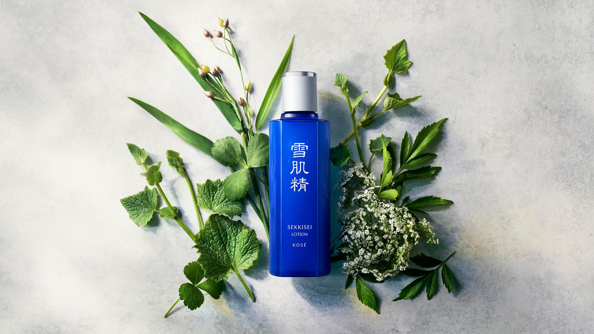 SEKKISEI Lotion bottle surrounded by green leaves and flowers on a light gray background
